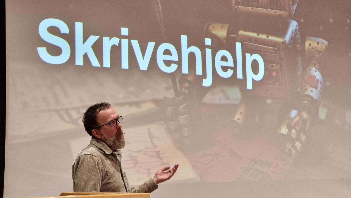 en mann med skjegg st&aring;r foran et publikum med et lysbilde med SKRIVEHJELP bak seg